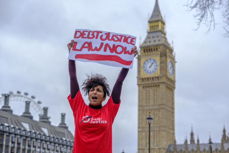 A person stands outside Parliament holding a sign reading “Debt Justice Law Now”, with the Elizabeth Tower behind them.