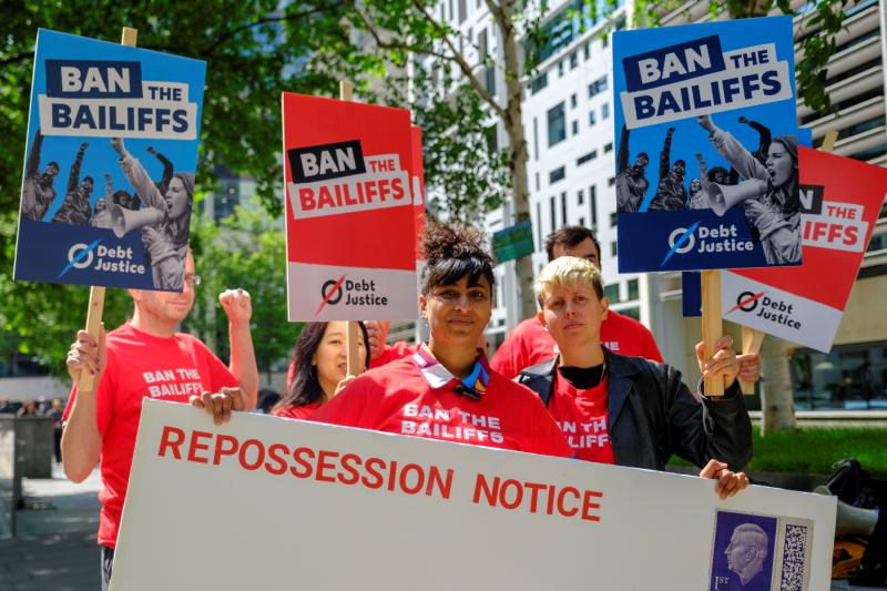 A group of protesters holdred and blue placards saying 'Ban the Bailiffs' in front of a giant envelope marked with 'Repossession Notice'