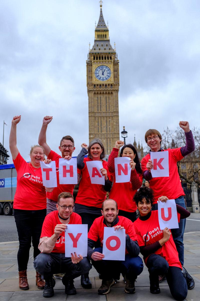 8 people in red Debt Justice t-shirts huddle together (5 on the top row and 3 on the bottom) holding red letters on a white background, spelling out the words 'Thank you'. They are smiling and the 5 at the back have their fists raised. 