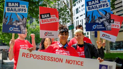 A group of protesters holdred and blue placards saying 'Ban the Bailiffs' in front of a giant envelope marked with 'Repossession Notice'